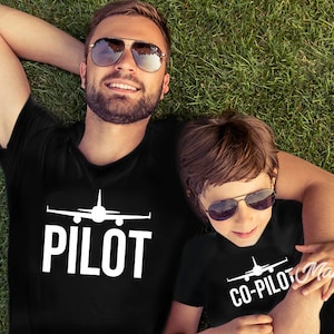May include: Two people wearing black t-shirts with white airplane graphics and text. The adult shirt says "PILOT" and the child's shirt says "CO-PILOT".
