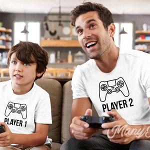 Puede incluir: Dos personas con camisetas blancas con un gr&aacute;fico de mando de juego negro y el texto "Player 1" y "Player 2". Est&aacute;n sosteniendo mandos de videojuegos y mirando la pantalla.