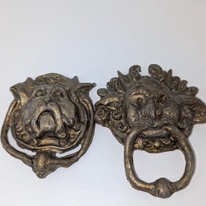 May include: Two antique-style door knockers. One features a bulldog face with a ring for knocking, and the other has a lion-like face with a ring. Both are bronze-coloured and have intricate details.