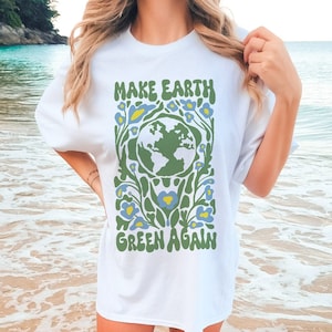 Op de afbeelding: Wit T-shirt met een groene en blauwe afbeelding van de aarde omringd door bloemen. De tekst "MAKE EARTH GREEN AGAIN" staat op het shirt gedrukt.