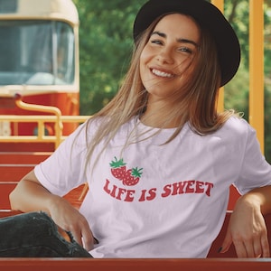 Op de afbeelding: Een vrouw draagt een wit T-shirt met een rode aardbei-afbeelding en de tekst "Life is Sweet". Ze zit op een bank met een rode en gele tram op de achtergrond.