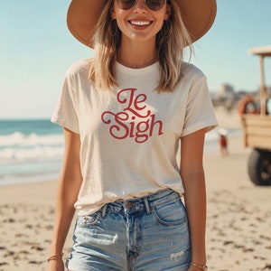 May include: A woman wearing a white t-shirt with the text "Le Sigh" in red lettering. She is wearing a brown hat, sunglasses, and denim shorts. She is standing on a beach.