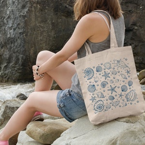 May include: A woman sitting on rocks by the ocean, holding a beige tote bag with a blue and white seashell design.