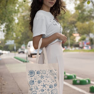 May include: A woman wearing a white t-shirt and a pink skirt is carrying a white tote bag with a blue seashell pattern. The bag has a variety of seashells, starfish, and other ocean-themed designs.