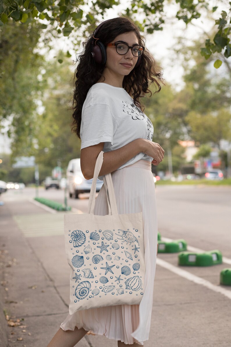 May include: A woman wearing a white t-shirt and a pink skirt is carrying a white tote bag with a blue seashell pattern. The bag has a variety of seashells, starfish, and other ocean-themed designs.