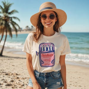 May include: A woman wearing a cream t-shirt featuring a pink and blue sardine can graphic, with the text "Sardines Introverts in a Can" and "Wild Caught". She wears a straw hat and sunglasses on a beach, with the sea in the background.