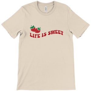 Op de afbeelding: Een beige T-shirt met een rode afbeelding van twee aardbeien en de tekst "LIFE IS SWEET" in rood.