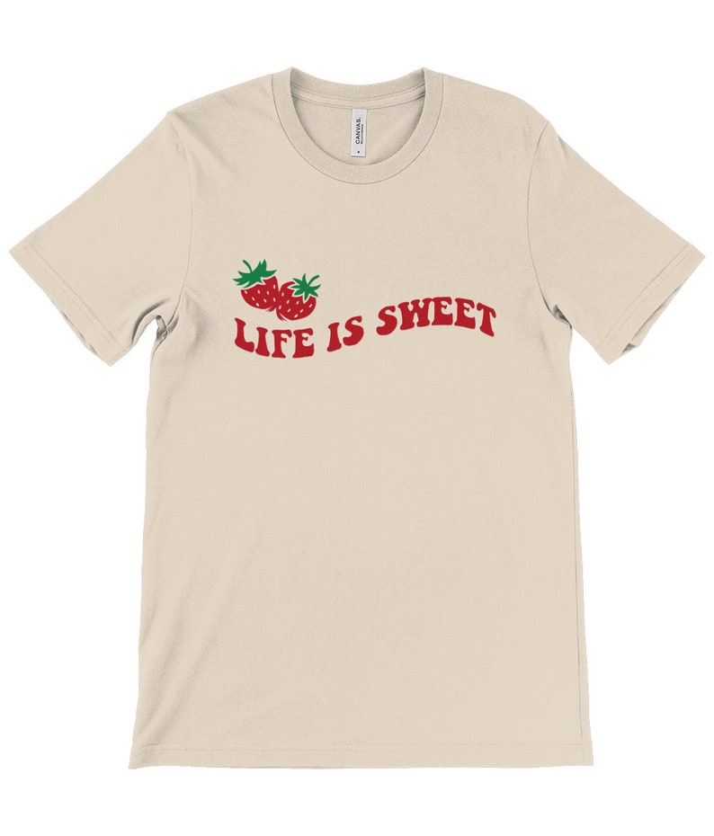 Op de afbeelding: Een beige T-shirt met een rode afbeelding van twee aardbeien en de tekst "LIFE IS SWEET" in rood.