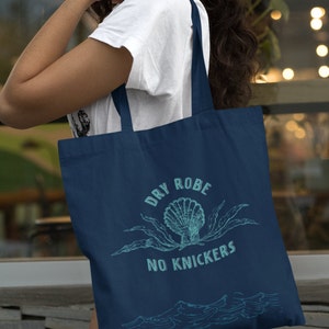 Op de afbeelding: Een donkerblauwe tote bag met een wit grafisch ontwerp met de tekst "Dry Robe No Knickers" met een schelp- en zeewierillustratie.