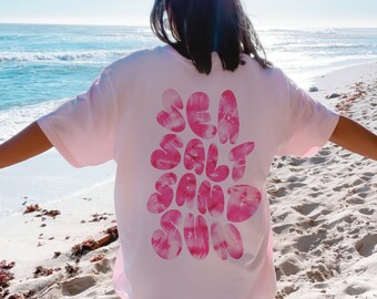 Può includere: Una maglietta rosa tie-dye con le parole "SEA SALT SAND SUN" in rosa sul retro. La maglietta è indossata da una persona in piedi su una spiaggia.