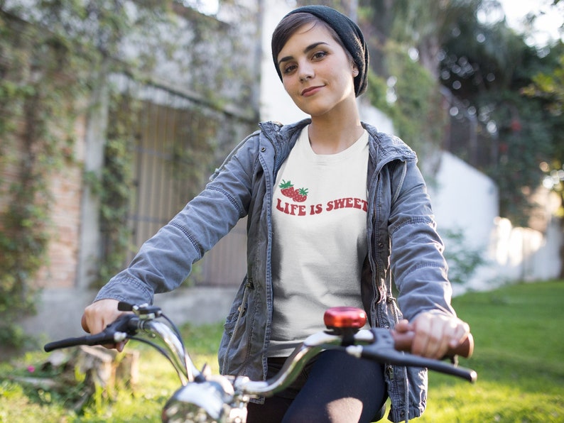 Op de afbeelding: Een wit T-shirt met een rode aardbei-afbeelding en de tekst "Life is Sweet". De persoon die het shirt draagt, fietst.