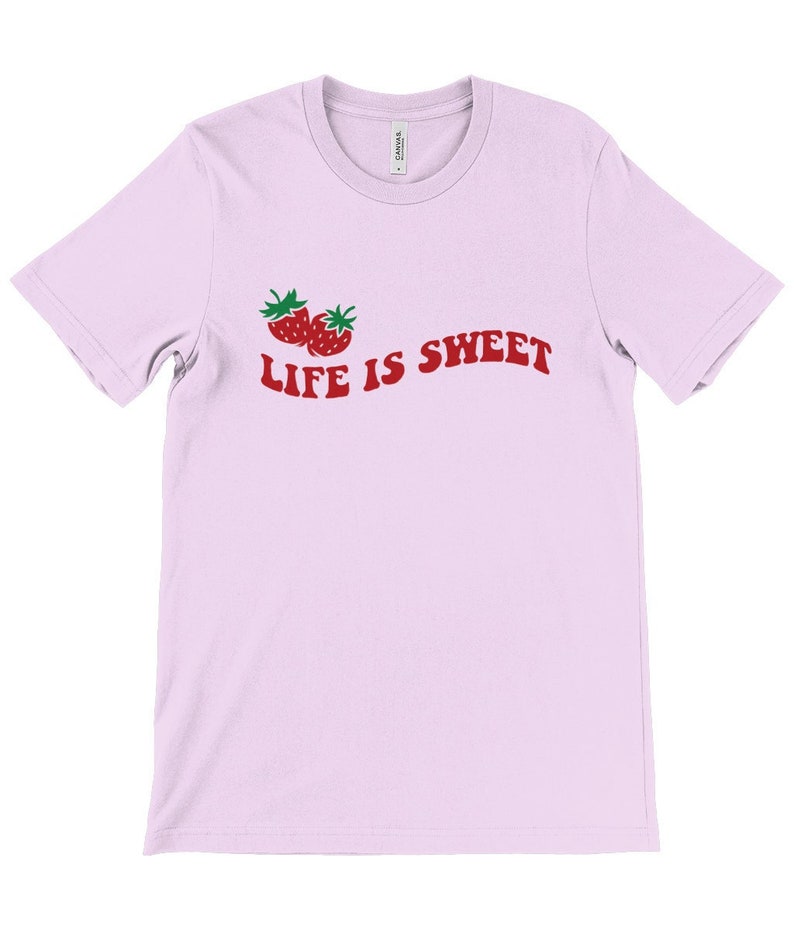 Op de afbeelding: Een lichtroze T-shirt met een rode afbeelding van twee aardbeien en de tekst "LIFE IS SWEET".