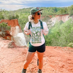 Op de afbeelding: Een persoon draagt een wit T-shirt met de tekst "Make Earth Green Again" en een groene afbeelding van de aarde omringd door bloemen. Ze draagt een zwarte broek, een groene baseballpet en een zonnebril. De persoon staat op een rood zandpad met uitzicht op een canyon op de achtergrond.