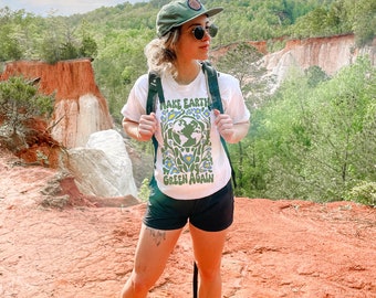 Puede incluir: Una persona lleva una camiseta blanca con el texto "Make Earth Green Again" y un gráfico verde de la Tierra rodeado de flores. Lleva pantalones cortos negros, una gorra de béisbol verde y gafas de sol. La persona está de pie en un camino de tierra roja con vista a un cañón en el fondo.
