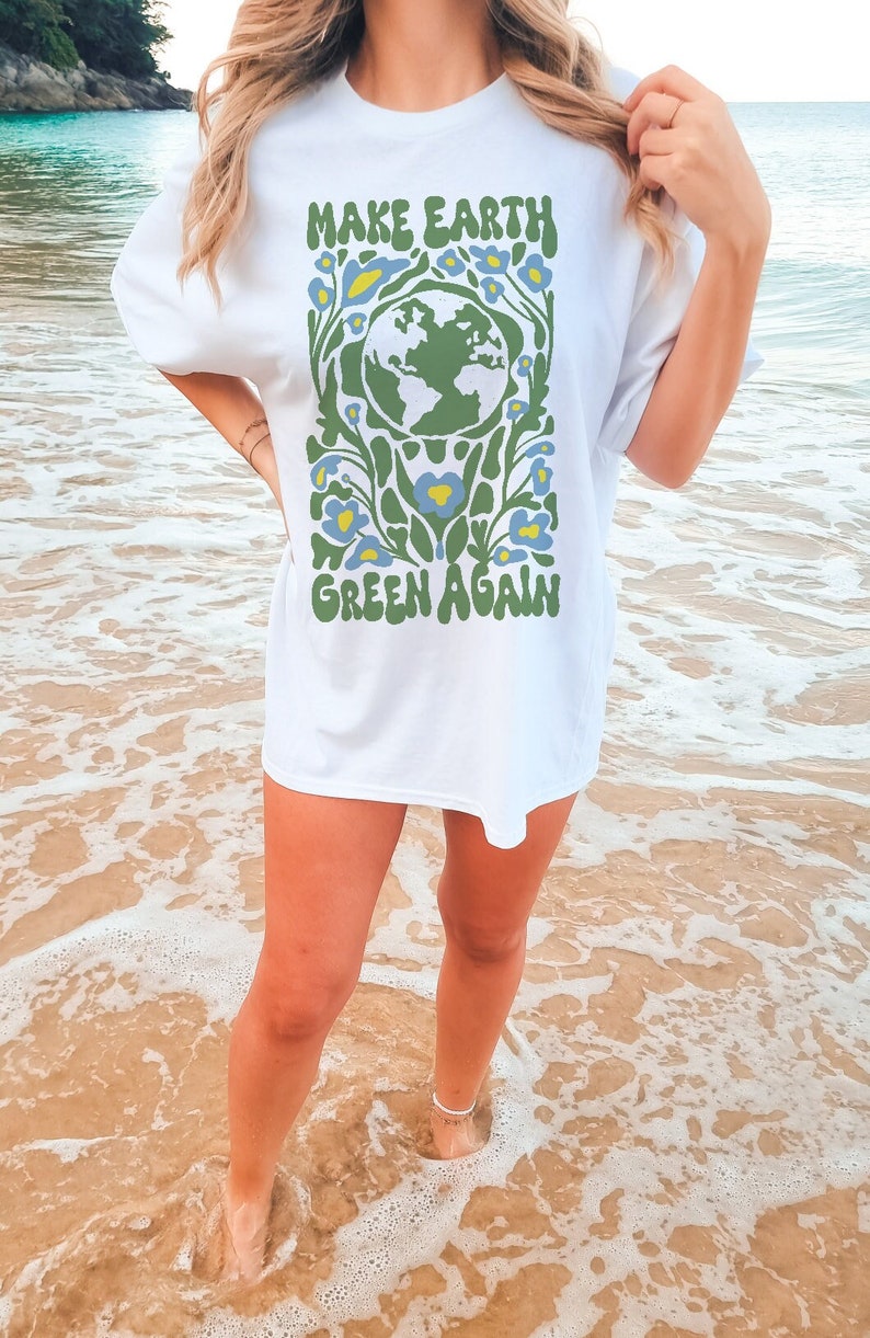 Op de afbeelding: Wit T-shirt met een groene en blauwe afbeelding van de aarde omringd door bloemen. De tekst "MAKE EARTH GREEN AGAIN" staat op het shirt gedrukt.