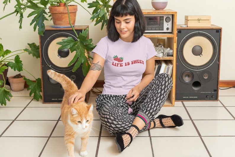 Op de afbeelding: Een vrouw draagt een lichtroze T-shirt met de tekst "Life is Sweet" en een aardbeienafbeelding en zit op de vloer met een kat. Ze draagt een zwart-wit geblokte broek en zwarte schoenen.