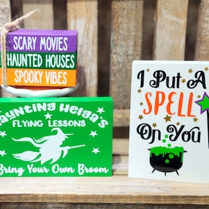 May include: Three wooden signs with Halloween themes. The first sign is a stack of three blocks with the text "Scary Movies", "Haunted Houses", and "Spooky Vibes". The second sign is green with white text that reads "Haunting Helga's Flying Lessons Bring Your Own Broom". The third sign is white with black text that reads "I Put A SPELL On You" with a green and purple star and a black cauldron with green liquid.