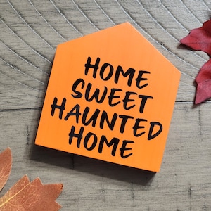 May include: Orange wooden sign with the text "Home Sweet Haunted Home" in black lettering.