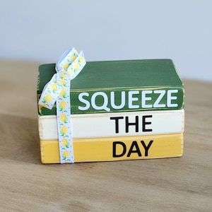 May include: A stack of three wooden blocks painted in green, cream, and yellow. The blocks are tied together with a ribbon printed with yellow pineapples. The words "SQUEEZE THE DAY" are painted on the blocks in black.