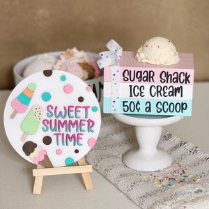 May include: A decorative ice cream-themed sign with the text "Sweet Summer Time" on a white circular board, and a wooden easel. A stack of colorful blocks reads "Sugar Shack Ice Cream 50¢ a Scoop" topped with a scoop of ice cream.