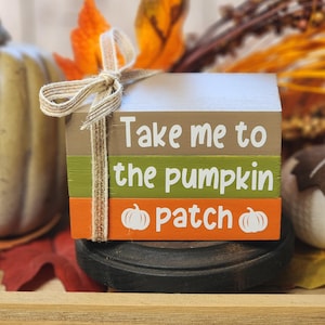 May include: A decorative wooden block set with the text "Take me to the pumpkin patch" in white. The blocks are painted in shades of brown, green, and orange, and tied with twine. The set is a fall-themed decoration.