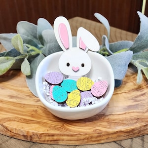 May include: A white ceramic bowl with a bunny rabbit cutout and pink ears. The bowl is filled with pastel colored wooden Easter eggs.