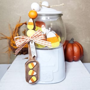 May include: White candy jar with a glass top filled with candy corn. The jar has a wooden spoon with the words "Candy Corn" hanging from it. The jar is decorated with a white and orange bow and a string of white and orange beads.