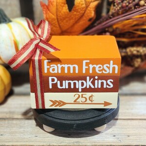 May include: A three-tiered wooden block sign with a red and white striped ribbon. The top block is orange and says "Farm Fresh Pumpkins". The middle block is brown and says "Pumpkins". The bottom block is cream and says "25¢" with an arrow pointing to the right.