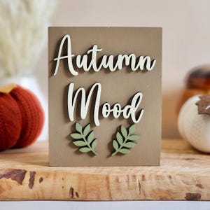 May include: A rectangular wooden sign with a light brown finish featuring the white 3D text "Autumn Mood" in a cursive script. Below the text are two small, light green wooden leaf cutouts centered on the lower portion of the sign.