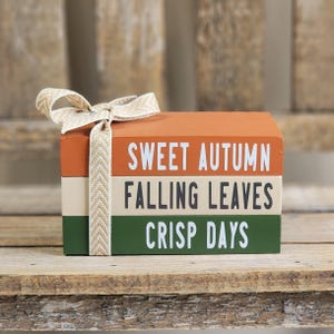 May include: A stack of three rectangular wooden blocks tied with a beige herringbone ribbon. The top block is orange with white text reading SWEET AUTUMN, the middle is cream with black text reading FALLING LEAVES, and the bottom is green with white text reading CRISP DAYS.