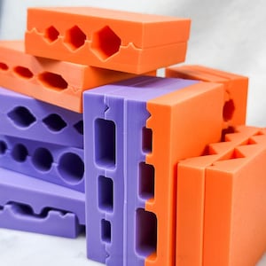 Puede incluir: Un surtido de moldes geométricos coloridos, en naranja y morado. Los moldes tienen varias formas, incluyendo hexágonos, círculos y recortes rectangulares. Las piezas están apiladas, mostrando su diseño entrelazado y colores vibrantes.