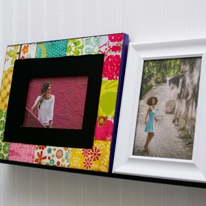 May include: Two picture frames with black and white frames, one with a colorful patchwork design and the other with a white frame. Both frames are hanging on a black shelf.