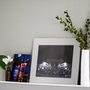 May include: A white shelf with two framed photographs and a white vase with green leaves. The photograph on the left shows a city street at night with colorful lights. The photograph on the right shows a car driving at night with its headlights on.