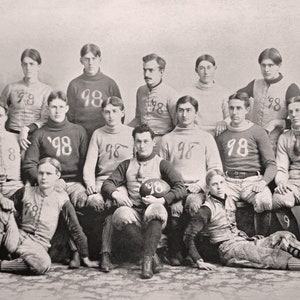 以下が含まれることがあります： 1898年のフットボールチームの白黒写真。チームは98番のユニフォームを着ています。選手たちはグループで立っており、一部は座っており、1人の選手がフットボールを持っています。
