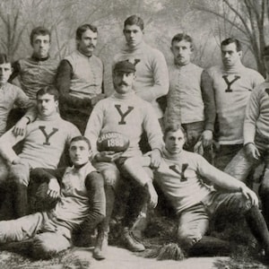 May include: A black and white photo of a group of men in athletic uniforms. The men are standing and sitting in a row. They are all wearing jerseys with the letter "Y" on them. The men are holding a football and a banner that says "Champions 1888".