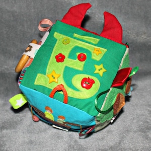 Può includere: Un cubo di tessuto multicolore con una grande lettera 'E' in feltro verde su un lato. Il cubo ha diverse texture e caratteristiche, tra cui bottoni, una mela rossa e una stella. È un giocattolo morbido e interattivo per neonati e bambini piccoli.