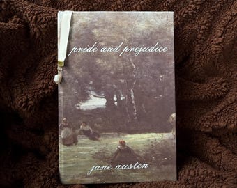 May include: A hardcover book titled Pride and Prejudice by Jane Austen with a cover featuring a landscape painting of figures in a field. A white ribbon bookmark with a small white bead charm is attached to the top edge of the book cover.