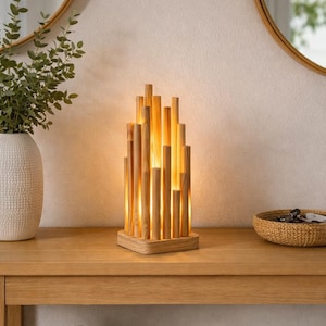 Puede incluir: Lámpara de mesa de madera con un diseño único. La lámpara presenta varillas verticales de madera de diferentes alturas, creando un brillo cálido. La lámpara se asienta sobre una base cuadrada de madera. La estética general es natural y moderna. Dimensiones: aprox. 20 cm.
