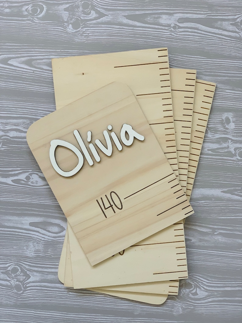 Peut inclure: Un tableau de croissance en bois avec une plaque nominative indiquant "Olivia" et une ligne de mesure marqu&eacute;e "140".