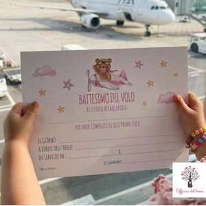 Può includere: Un bambino tiene in mano un certificato con la scritta "BATTESIMO DEL VOLO" e un orsetto dei cartoni animati su un aereo rosa. Il certificato è tenuto davanti a un aereo ITA Airways in aeroporto.