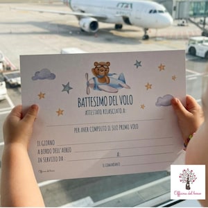 Può includere: Un bambino tiene in mano un certificato con la scritta "BATTESIMO DEL VOLO" e un orsetto dei cartoni animati che pilota un aereo. Un aereo è visibile sullo sfondo. Il certificato è per un primo volo.