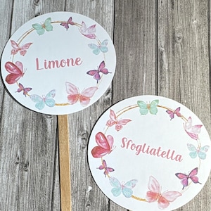 Può includere: Due cartelli rotondi bianchi su bastoncini di legno, ciascuno con un disegno di farfalle rosa, blu e verdi. Le farfalle circondano il bordo del cerchio. Un cartello dice "Limone", l'altro "Sfogliatella".