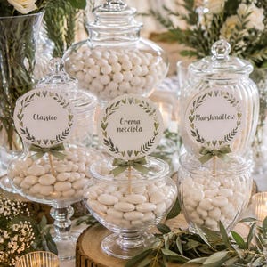 Può includere: Tre barattoli in vetro trasparente riempiti con mandorle confitate bianche. Ogni barattolo presenta un cartellino rotondo bianco con un disegno di corona di foglie verdi e il testo Classico, Crema nocciola o Marshmallow, legato con un nastro verde.