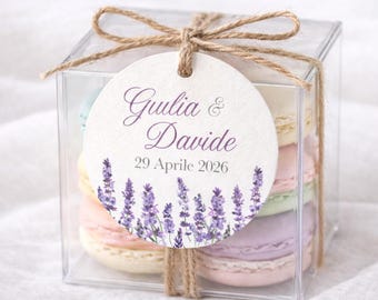 May include: A clear, square box tied with twine, containing colorful macarons. A round tag with the names "Guilia & Davide" and the date "29 Aprile 2026" is attached. Lavender sprigs decorate the bottom of the tag.