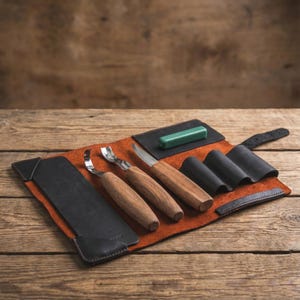 Pode incluir: Um conjunto de ferramentas de entalhe em madeira com cabos de madeira e lâminas de metal, apresentado em um rolo de couro marrom. O conjunto inclui uma goiva curva, uma faca reta e uma pedra de amolar.