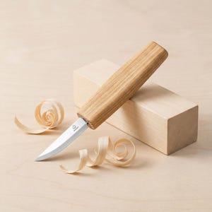 May include: A wood carving knife with a light-colored wooden handle and a silver blade. The knife is positioned on a light-colored wooden surface, with wood shavings and a wooden block in the background. The knife has a small logo on the blade.