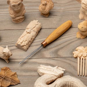 May include: A wood carving tool with a light brown wooden handle and a copper ferrule, positioned next to several small hand-carved wooden figures including a monkey, a turkey, a green man, a rabbit, a dragon, and a decorative hair comb.