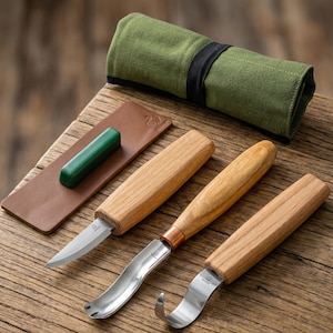 May include: A wood carving set with three knives, a sharpening stone, and a leather strop. The knives have wooden handles and stainless steel blades. A green canvas roll holds the tools. The set is displayed on a wooden surface.