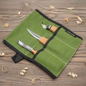 May include: A green canvas tool roll containing three wood carving knives. The knives have wooden handles and stainless steel blades. The tool roll is open, displaying the tools, and is surrounded by wood shavings.