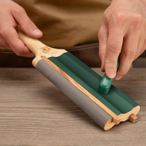May include: A wooden and green sharpening tool with a grey base. A small green stone is being used on the tool. The handle is light-coloured wood, and the tool has a decorative design.